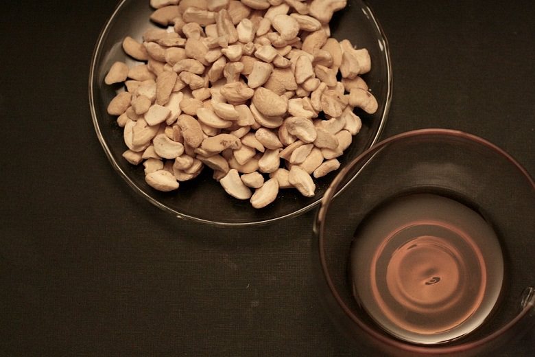 Brokkoli-Cashew Salat mit Mandarinen - Cashewkerne und Olivenöl in Glasschüsseln vor dunklem Hintergrund.